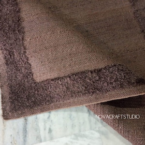 May include: Close-up of a brown rug with a textured weave and a darker, fringed border. The rug is folded, revealing the underside. The image is well-lit, showcasing the details of the textile.