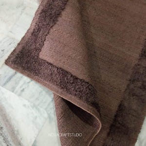 May include: A folded brown woven textile with a textured border. The textile has a rectangular shape and appears to be made of a natural fiber. The brand name "NOVACRAFTSTUDIO" is visible in the image.