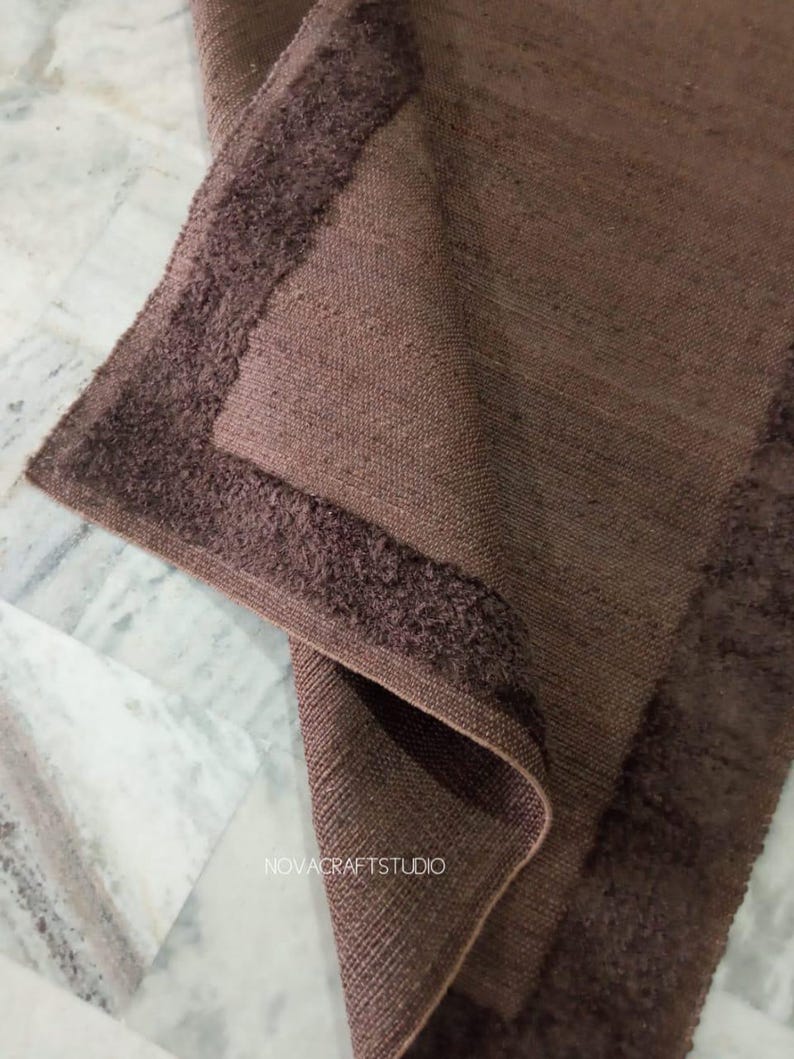 May include: A folded brown woven textile with a textured border. The textile has a rectangular shape and appears to be made of a natural fiber. The brand name "NOVACRAFTSTUDIO" is visible in the image.