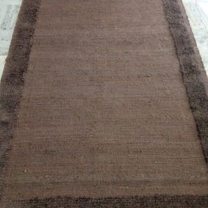 May include: A rectangular brown rug with a darker brown border. The rug has a woven texture and a plush border. The rug is a neutral color and would complement many home decor styles. The rug is suitable for a living room or bedroom.