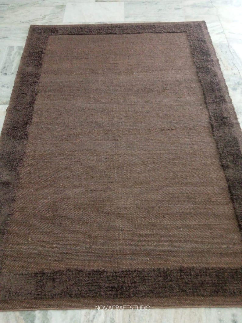 May include: A rectangular brown rug with a darker brown border. The rug has a woven texture and a plush border. The rug is a neutral color and would complement many home decor styles. The rug is suitable for a living room or bedroom.