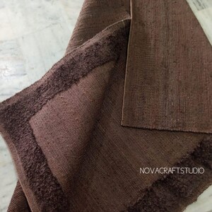 May include: A brown rug with a textured surface and a darker brown, fringed border. The rug is folded over, revealing the underside. The text "NOVACRAFTSTUDIO" is visible in the lower right corner.