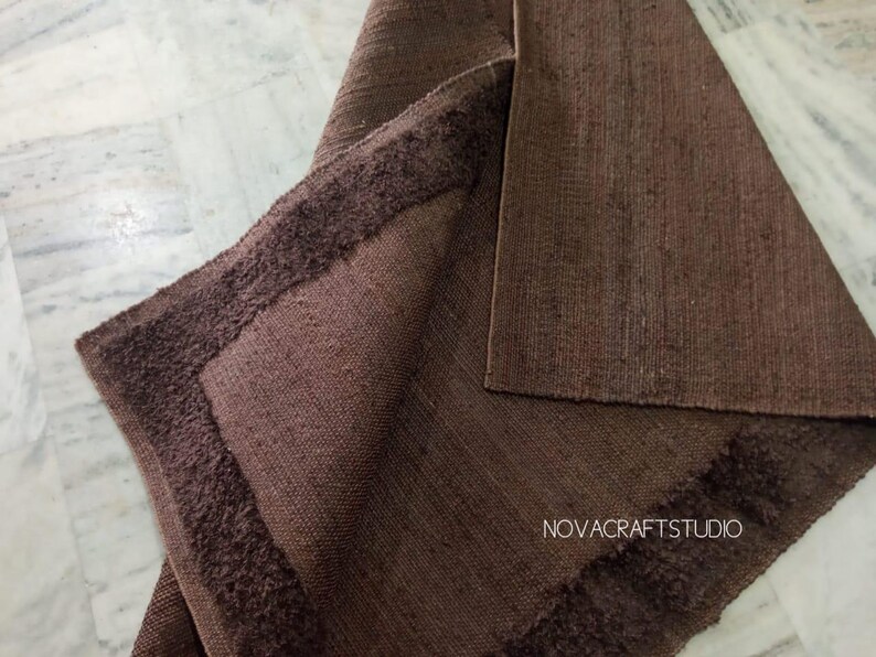 May include: A brown rug with a textured surface and a darker brown, fringed border. The rug is folded over, revealing the underside. The text "NOVACRAFTSTUDIO" is visible in the lower right corner.