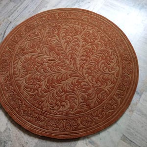 May include: A round, rust-colored rug with an intricate floral pattern. The rug features a central design of stylized leaves and flowers, surrounded by a detailed border. The rug is made of a soft material and is suitable for home decor.