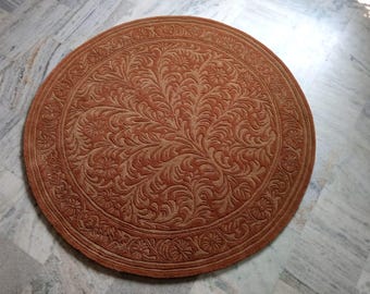 May include: A round, rust-colored rug with an intricate floral pattern. The rug features a central design of stylized leaves and flowers, surrounded by a detailed border. The rug is made of a soft material and is suitable for home decor.