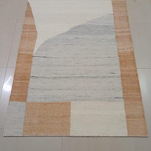 May include: A rectangular area rug featuring a woven texture with a color palette of tan, off-white, and light gray. The design consists of a wide tan border framing a central section with large, abstract geometric shapes in white and gray.