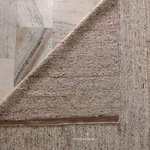 Op de afbeelding: Een beige en bruin tapijt met zichtbare textuur, gedeeltelijk opgevouwen om de onderkant te laten zien. Het tapijt heeft een zichtbare rand en een gespikkelde uitstraling. De achtergrond is een marmerachtig oppervlak.