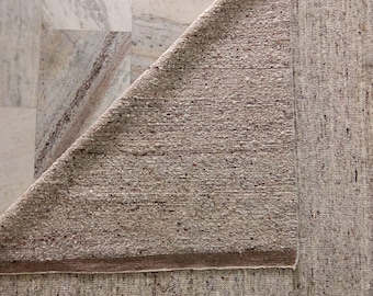 May include: A neutral-toned rug with a textured surface, partially folded to reveal the underside. The rug's color is a blend of beige and brown, with a visible border. The background features a marble-like surface.