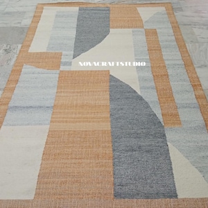 May include: Rectangular area rug with a geometric design. The rug features a combination of colors, including beige, gray, and white. The design incorporates curved and straight lines, creating an abstract pattern. The rug is made of a woven material.
