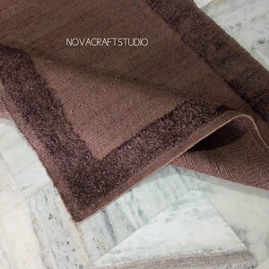 May include: A folded brown rug with a textured weave and a darker, fringed border. The rug's corner is visible, displaying the contrasting textures and colors. The text "NOVACRAFTSTUDIO" is visible in the image.
