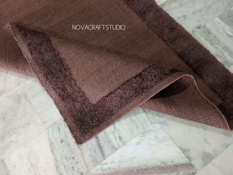 May include: A folded brown rug with a textured weave and a darker, fringed border. The rug's corner is visible, displaying the contrasting textures and colors. The text "NOVACRAFTSTUDIO" is visible in the image.