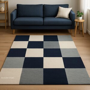 Puede incluir: Una alfombra rectangular con un gran diseño de tablero de ajedrez en azul marino, gris claro y blanco roto. La alfombra está colocada sobre un suelo de madera frente a un sofá azul marino. La alfombra tiene el texto "NOVACRAFTSTUDIO".