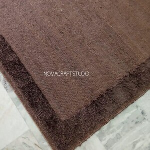 May include: A brown rectangular rug with a darker brown border. The rug has a textured appearance and is placed on a marble surface. The text "NOVACRAFTSTUDIO" is visible on the rug.