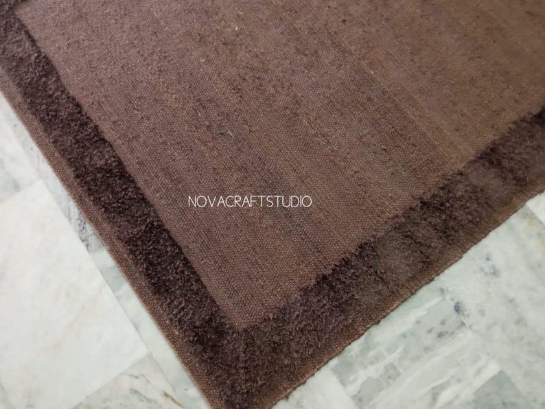 May include: A brown rectangular rug with a darker brown border. The rug has a textured appearance and is placed on a marble surface. The text "NOVACRAFTSTUDIO" is visible on the rug.
