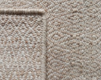 May include: Close-up of a beige and tan woven rug. The rug has a textured surface with a diamond pattern. The edge of the rug is visible, showing a finished border. The rug is made of a natural fiber.