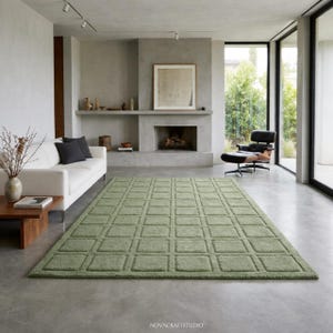 May include: A large, rectangular, sage green rug with a repeating square pattern dominates the room. The rug is placed in front of a white sofa and a fireplace. The room has a modern design with large windows and a minimalist aesthetic.