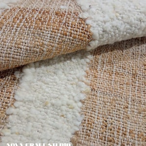 May include: Close-up of a woven rug with a textured design. The rug features alternating sections of beige and off-white, with a looped, shaggy texture in the white areas. The words "NOVA CRAFT STUDIO" are printed on the rug.