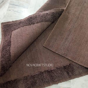 May include: A folded brown rug with a textured surface and a darker, fringed border. The rug's corner is turned up, revealing the underside. The text "NOVACRAFTSTUDIO" is visible on the rug.