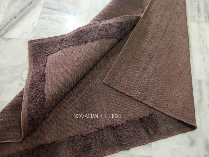 May include: A folded brown rug with a textured surface and a darker, fringed border. The rug's corner is turned up, revealing the underside. The text "NOVACRAFTSTUDIO" is visible on the rug.