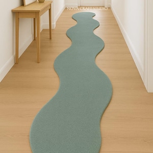 May include: A teal, wavy-shaped rug in a hallway. The rug has a soft, textured appearance and a unique, organic shape. The hallway has light wood flooring and white walls. A wooden table with a framed picture and a vase of yellow flowers is visible.