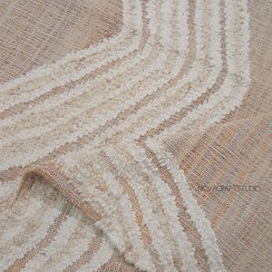 May include: Close-up of a beige and cream-colored textile with a textured, looped pattern. The design features curved, off-white stripes against a neutral background. The fabric is folded, revealing its layered construction.