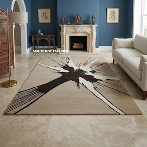 May include: A rectangular area rug featuring a large, abstract ink-blot style design in dark brown and cream against a light tan background. The pattern radiates from the center with sharp, elongated points and fine, linear brushstroke details.