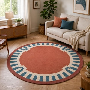 May include: A round rug with a terracotta center and a border featuring a geometric design in cream and teal. The rug is placed in a living room setting with a sofa, pillows, and a wooden cabinet.