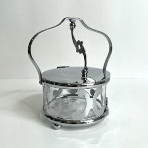 May include: A circular glass serving dish encased in a polished silver-tone metal frame with a decorative scrollwork pattern. The container features a matching silver-tone metal lid and a tall, arched handle attached to the top center.