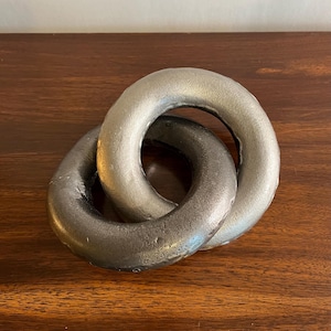 Puede incluir: Una escultura decorativa que consiste en dos anillos circulares entrelazados hechos de un material metálico con un acabado texturizado en gris plateado oscuro. Los anillos están conectados formando una cadena continua.