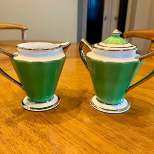 Op de afbeelding: Een vintage suiker- en roomstel in groen en wit. De stukken hebben een geometrisch ontwerp met zilveren randen en handvatten. De suikerpot heeft een bijpassend deksel met een gouden knop. De set staat op een houten tafel.