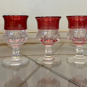 Puede incluir: Cinco copas de cristal transparente con bordes rojo rubí y cuencos de cristal tallado decorativos. Las copas tienen un tallo y una base transparentes. Las copas están dispuestas sobre una superficie reflectante.