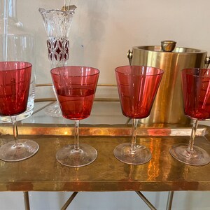 Puede incluir: Cuatro copas de vino tinto con un diseño geométrico están dispuestas sobre una bandeja de latón. Una cubitera dorada y una jarra de cristal transparente están en el fondo. Las copas tienen tallos largos y cuencos anchos e inclinados.