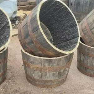 May include: Three weathered wooden barrels with metal bands. The barrels are grey with dark interiors and are stacked on top of each other. The barrels are outdoors on a concrete surface.