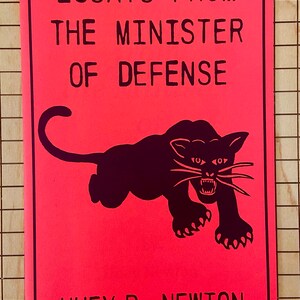 May include: A red book cover with black text that reads "Essays From The Minister Of Defense" and a black silhouette of a panther with a white face and white whiskers. The author's name, "Huey P. Newton", is printed in black at the bottom of the cover.