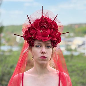 May include: A red floral headpiece with a veil. The headpiece is adorned with numerous red roses and small gold accents. The veil is a sheer red fabric. The person is wearing a red bra.