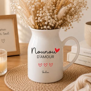 Peut inclure: Un pichet en céramique blanc avec une anse, rempli de fleurs séchées. Le pichet porte les mots "Nounou D'Amour" et trois cœurs rouges, avec le nom "Julia" en dessous. Une photo encadrée et une bougie sont en arrière-plan.