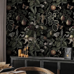 May include: Dark, moody interior scene featuring a black botanical wallpaper with leopards, fruit, and butterflies. A black table holds open books, a candle, and a coffee cup. A dark cabinet displays gold candlesticks and a gift bag.