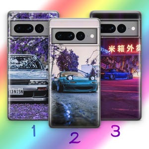 以下が含まれることがあります： 自動車デザインのスマートフォンケース3点。ケース1は紫色の木々の下にあるナンバープレート「13-NISN」の銀色の車。ケース2はリトラクタブルヘッドライトのターコイズブルーの車。ケース3はネオン街の青い車が描かれています。