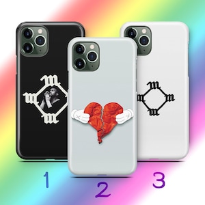 May include: Three phone cases with distinctive designs. A black case has a praying figure within a white emblem. A light grey case displays a broken heart held by cartoon hands. A white case has a white emblem.