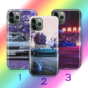 Puede incluir: Tres fundas de teléfono con diseños de coches. Una funda presenta un coche plateado con matrícula, otra un coche deportivo turquesa y la tercera muestra un coche azul bajo luces de neón. Cada funda tiene un borde negro.