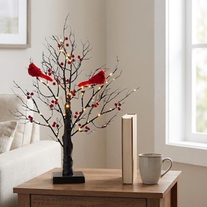 Könnte beinhalten: Ein dekorativer schwarzer Baum mit roten Beeren und zwei roten Kardinalvogel-Ornamenten. Der Baum ist mit kleinen weißen Lichtern beleuchtet und steht auf einem schwarzen quadratischen Sockel. Ein Buch und eine Tasse stehen auf dem Holztisch.