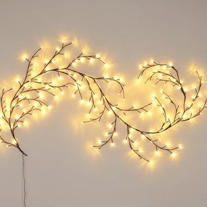 May include: A decorative, illuminated branch garland with warm white LED lights. The garland features dark brown branches with numerous small lights, creating a soft, glowing effect. The lights are evenly spaced along the branches, and a power cord is visible.