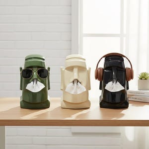 May include: Three Moai statue tissue box holders in green, cream, and black. The green Moai wears sunglasses, the black one has headphones, and all three dispense tissues from their mouths. They are displayed on a wooden table.