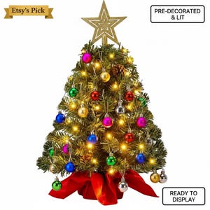 May include: A pre-decorated and lit miniature Christmas tree with a gold star topper. The tree is adorned with colorful ornaments and lights, and sits atop a red fabric base. The text "READY TO DISPLAY" is visible.