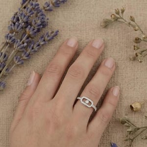 May include: A silver ring with a rectangular center design, encrusted with small, clear stones. A single, larger clear stone is set in the center of the rectangle. The ring is on a person's finger, with lavender and dried flowers in the background.