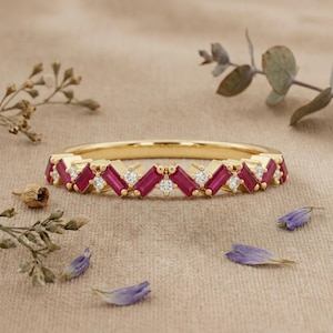 May include: A gold band ring featuring an alternating pattern of baguette-cut red rubies and small round-cut white diamonds. The stones are set in a continuous row along the top half of the polished yellow gold metal band.