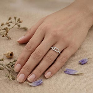 May include: A silver ring with a unique oval design, encrusted with small, sparkling stones. The ring is displayed on a hand, with a neutral background and scattered dried flowers and purple petals.