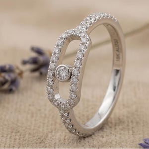 May include: A silver ring with a unique design. The ring features a central oval shape with a single, prominent diamond and is encrusted with smaller diamonds. The band is also adorned with small diamonds, creating a sparkling effect. The ring is set against a neutral background.