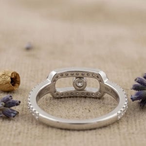May include: A silver ring with a rectangular center stone and a halo of smaller stones. The band is also adorned with small stones. The ring is set against a neutral background with hints of purple and brown.