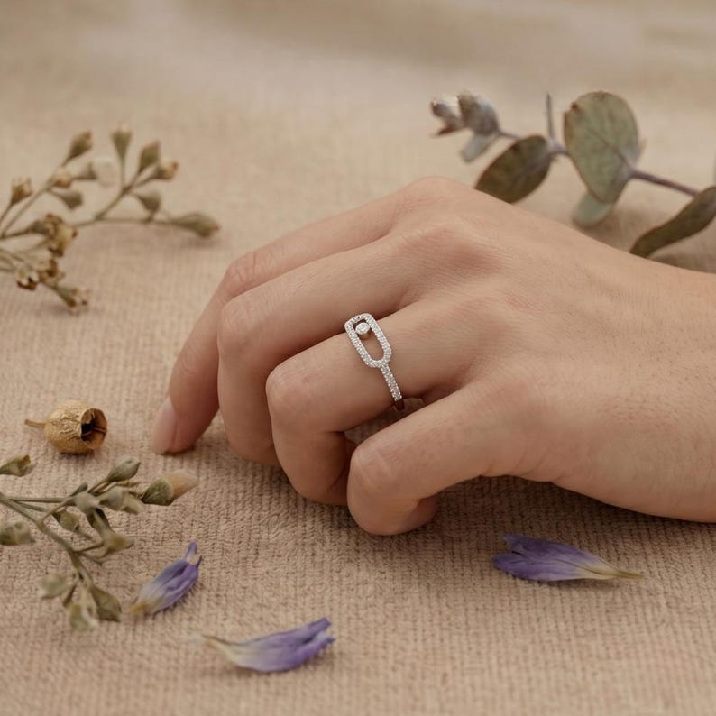 May include: A silver ring with a rectangular design and a small, round gemstone. The ring is set with small, sparkling stones along the band and the rectangular frame. The ring is on a person's finger.